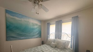 Een slaapkamer met een bed, een plafondventilator, een schilderij van een golf en een raam met gordijnen.