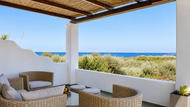 Uma área de estar coberta ao ar livre, com móveis de vime, uma mesa e vista para o mar.