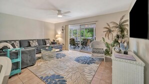 A living room with a sofa, coffee table, and a large floral rug. There is a ceiling fan, a window with a view of greenery, and a white wicker chair. - Your own Tropical Resort Heated Pool Pickleball (St. Petersburg)