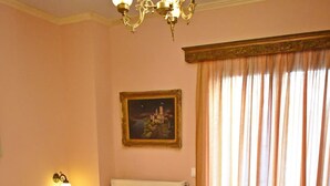 Una cama con una colcha amarilla, almohadas blancas y toallas enrolladas. Hay un cuadro en la pared, un radiador y una ventana con cortinas.