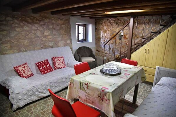 A cozy room with a stone wall, a white sofa with red cushions, a dining table set with plates and a bowl, and a staircase with a metal railing.