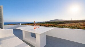 A white outdoor seating area with a table and chairs, a bottle, and two glasses, overlooking a coastal landscape. - Villa Therme Athen-sea View Jacuzzi Santorini Stay (Santorini)