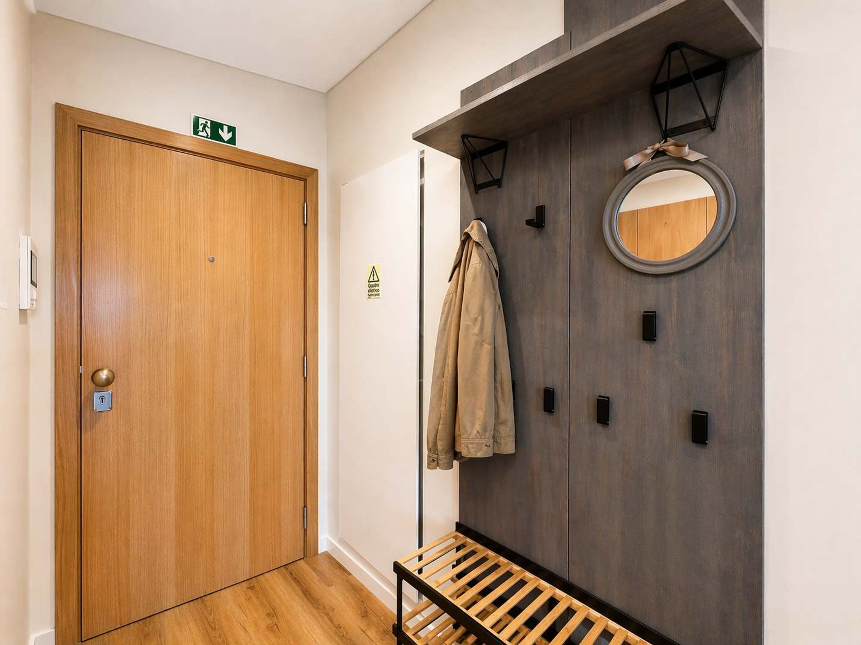 A hallway with a wooden door, an emergency exit sign, a coat rack with a jacket, a bench, and a wall-mounted mirror.
