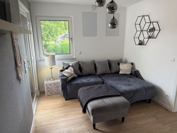 A modern bedroom with a grey sectional bed, a small bedside table with a lamp, a geometric wall decoration, and a view of greenery through a window.