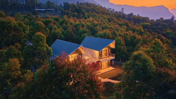Una casa moderna con techo plano, rodeada de un bosque denso y montañas al fondo.
