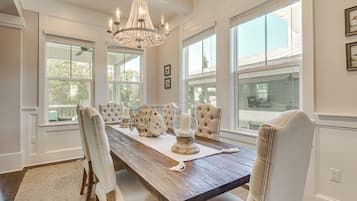 Un espace repas comprenant une table en bois, des chaises rembourrées, un lustre et de grandes fenêtres.