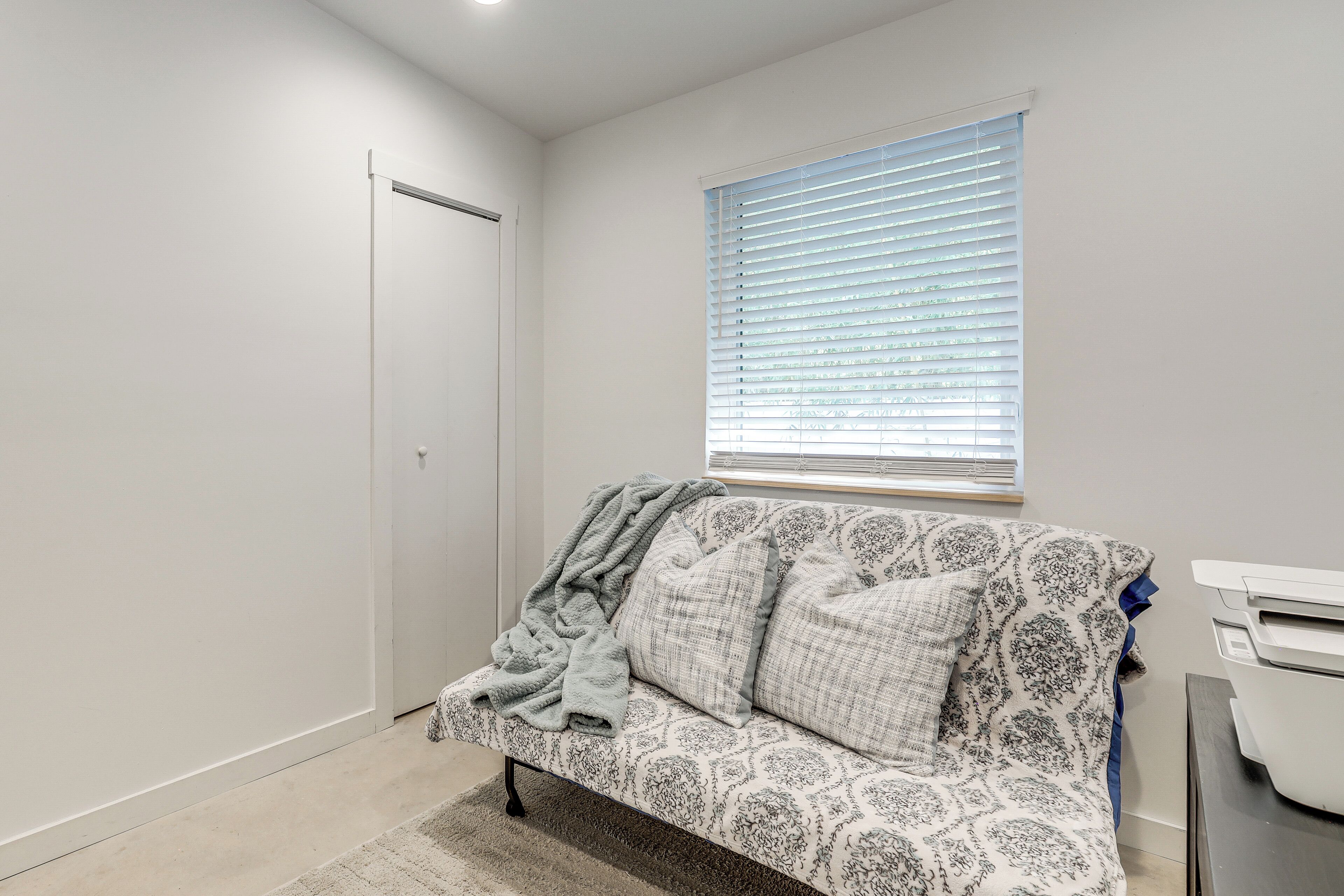 Una habitación con un sofá estampado, una ventana con persianas, una puerta e impresora sobre una mesa.