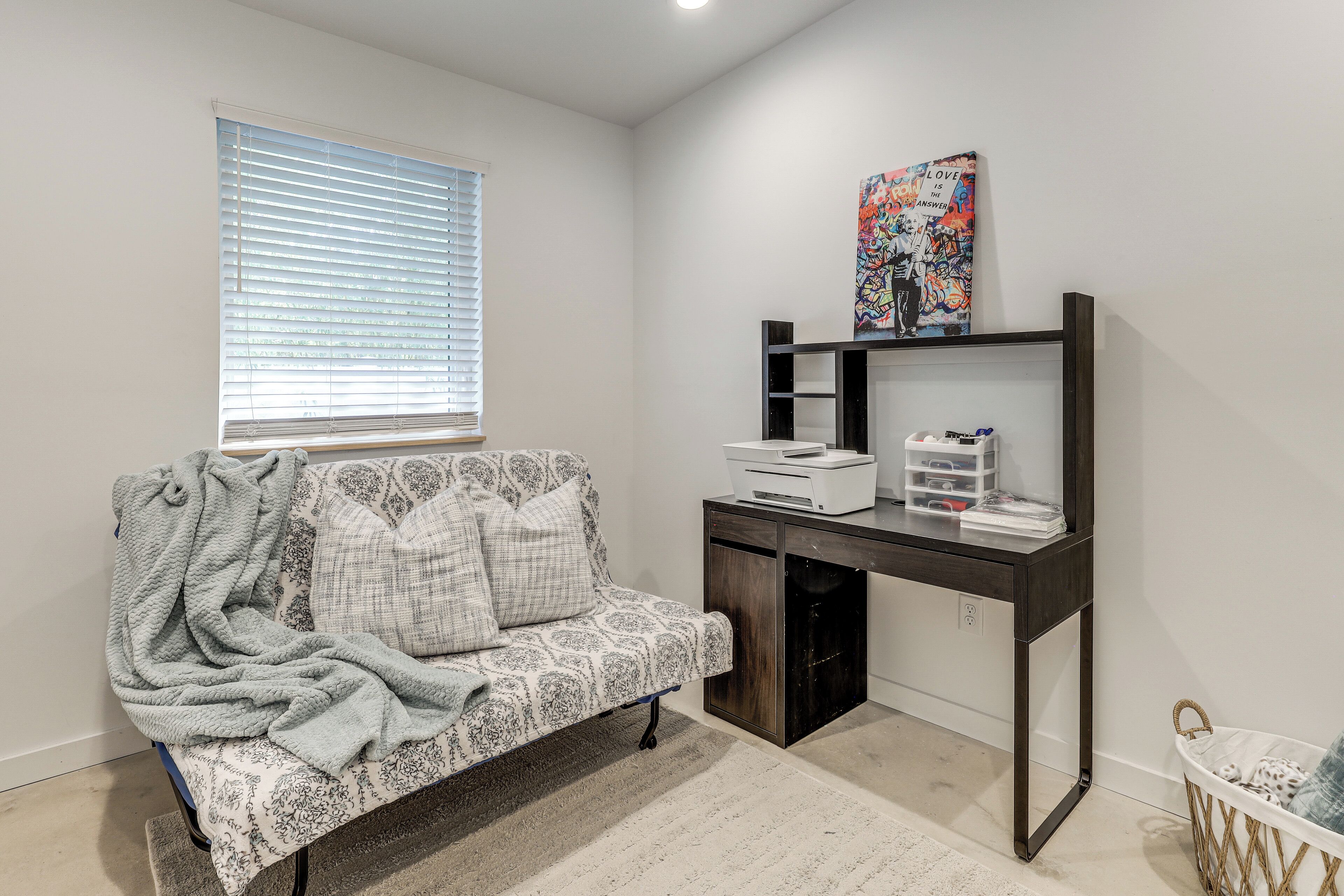 A room with a patterned sofa, a desk with storage, a window with blinds, and a wall-mounted artwork.