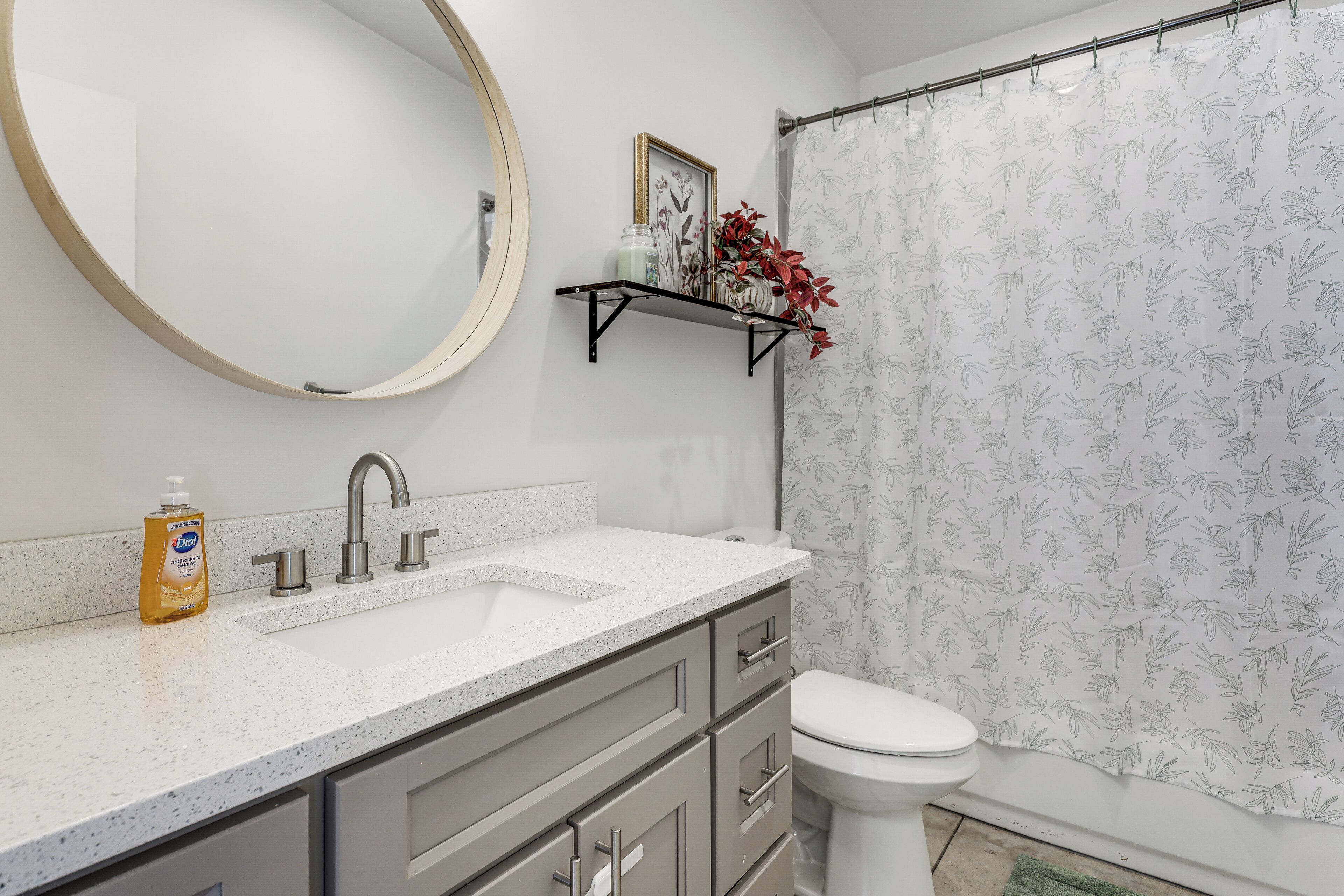 Ein Badezimmer mit rundem Spiegel, Waschbecken mit Wasserhahn, Toilette und einer Duschkabine mit Blumenmuster.