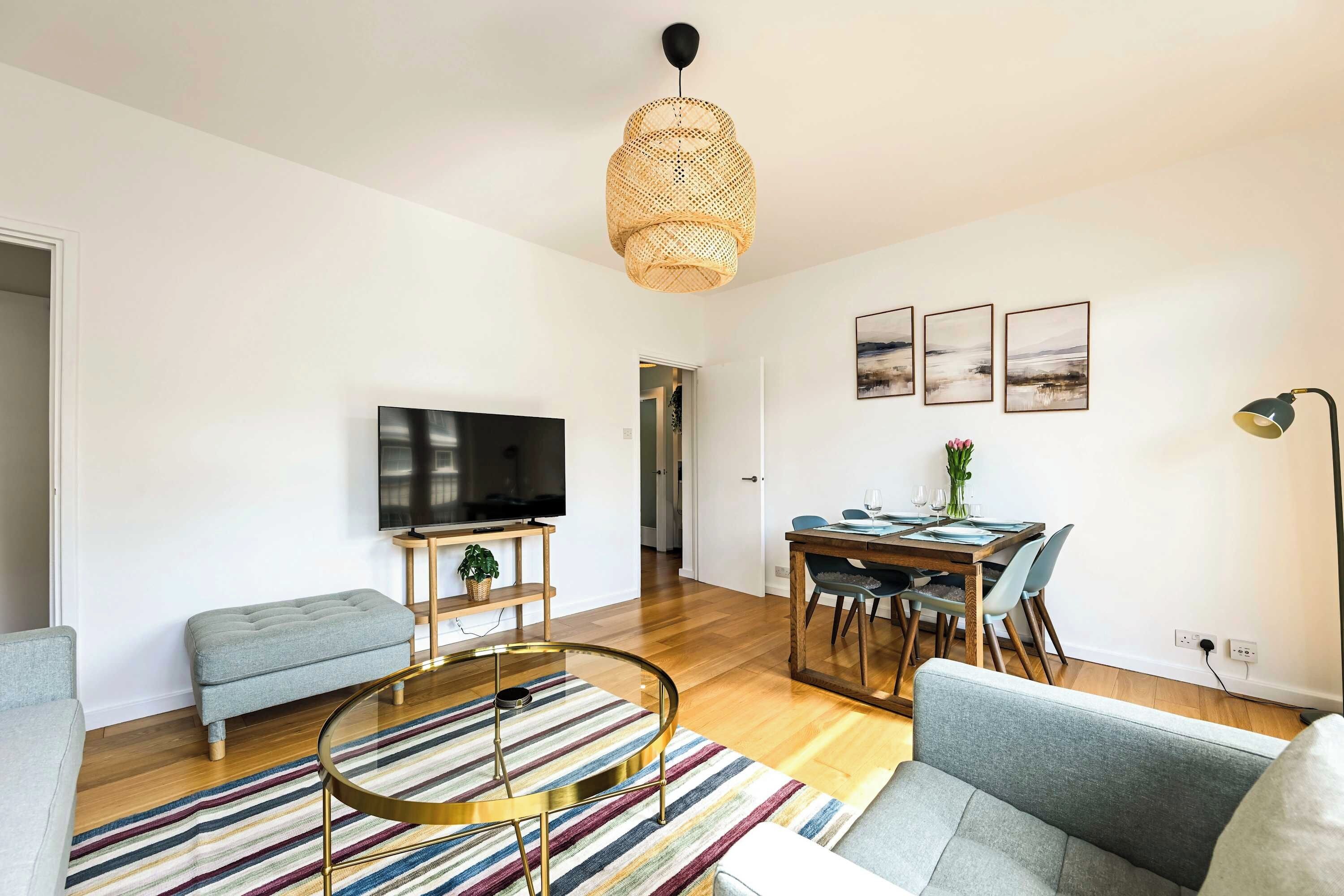 Ein modernes Wohnzimmer mit Flachbildfernseher, rundem Couchtisch, grauem Sofa, Teppich mit Streifen, einer Essecke mit Holztisch und Stühlen sowie einer hängenden Korbschwingerleuchte.