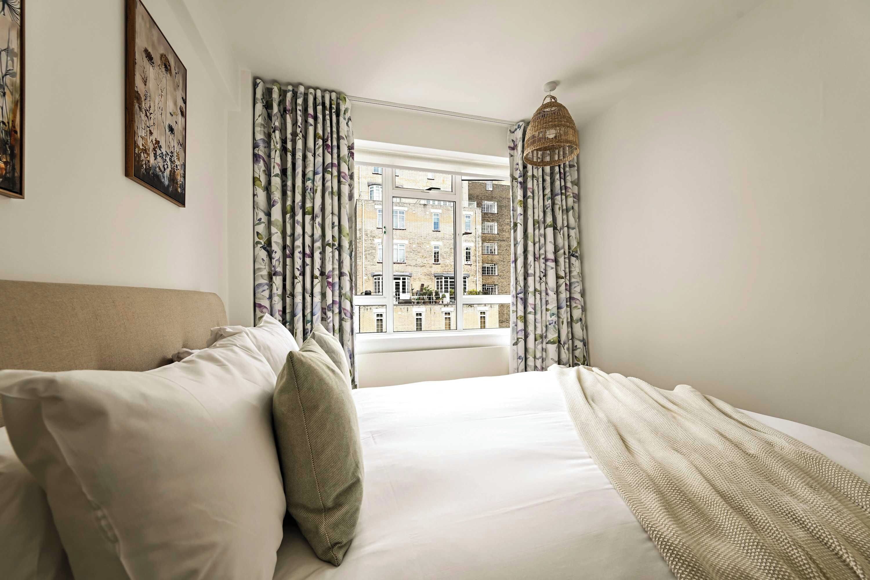 Une chambre à coucher avec un lit, des rideaux, un luminaire suspendu et une vue sur les bâtiments par la fenêtre.