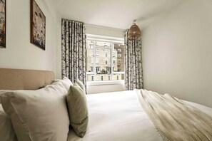A bedroom with a bed, curtains, a hanging light fixture, and a view of buildings through the window. - British Museum Apartment (Greater London)