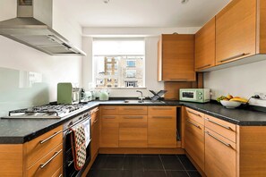 A modern kitchen with wooden cabinets, a gas stove, a microwave, and a window with a city view. - British Museum Apartment (Greater London)