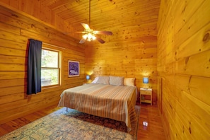 A wooden cabin bedroom with a bed, two bedside tables with lamps, a ceiling fan, a window with curtains, and a framed picture on the wall. - Butler Cabin w/ 2 Decks: Near Watauga Lake! (Mountain City)