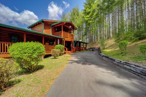 Une cabane en rondins à deux étages, dotée d’un balcon, entourée d’une zone boisée et accessible par une allée pavée.
