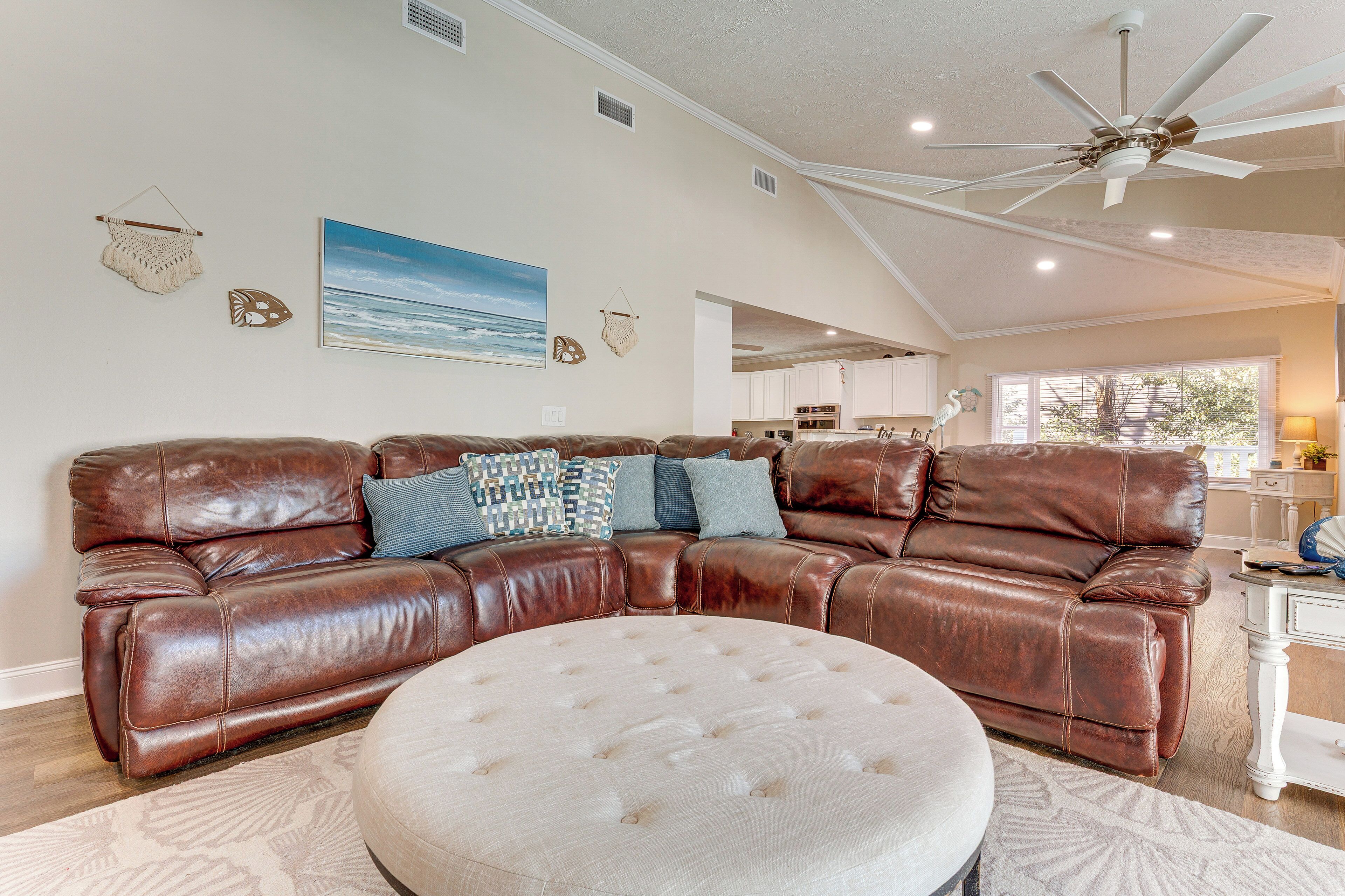 A spacious living room with a brown leather sectional sofa, a round ottoman, and a ceiling fan.