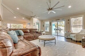 Ein geräumiges Wohnzimmer mit einem braunen Ledersofa, einem runden Hocker, Deckenventilatoren und großen Fenstern.