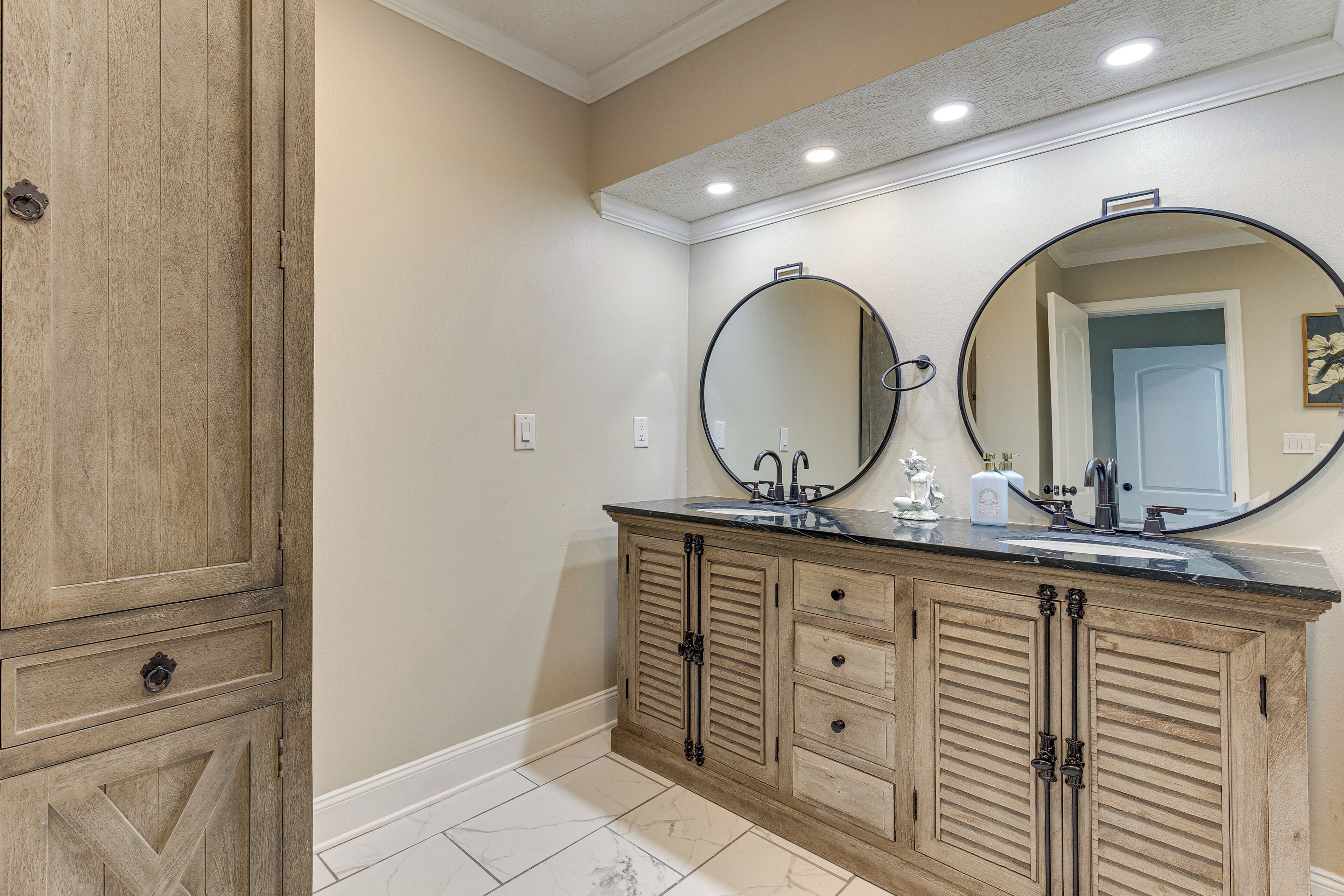 A bathroom with a wooden vanity, two round mirrors, a sink, and a door leading to another room.