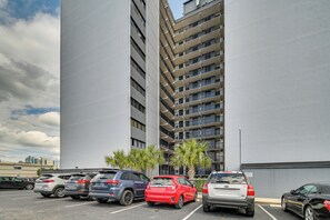 A parking lot with several cars, a tall building with multiple balconies, and some palm trees. - Walk to Family Kingdom! Condo w/ Pool + Balcony (Myrtle Beach)