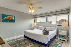 Ein Schlafzimmer mit Bett, Deckenventilator, Fenster mit Jalousien und Blick auf die Stadt.