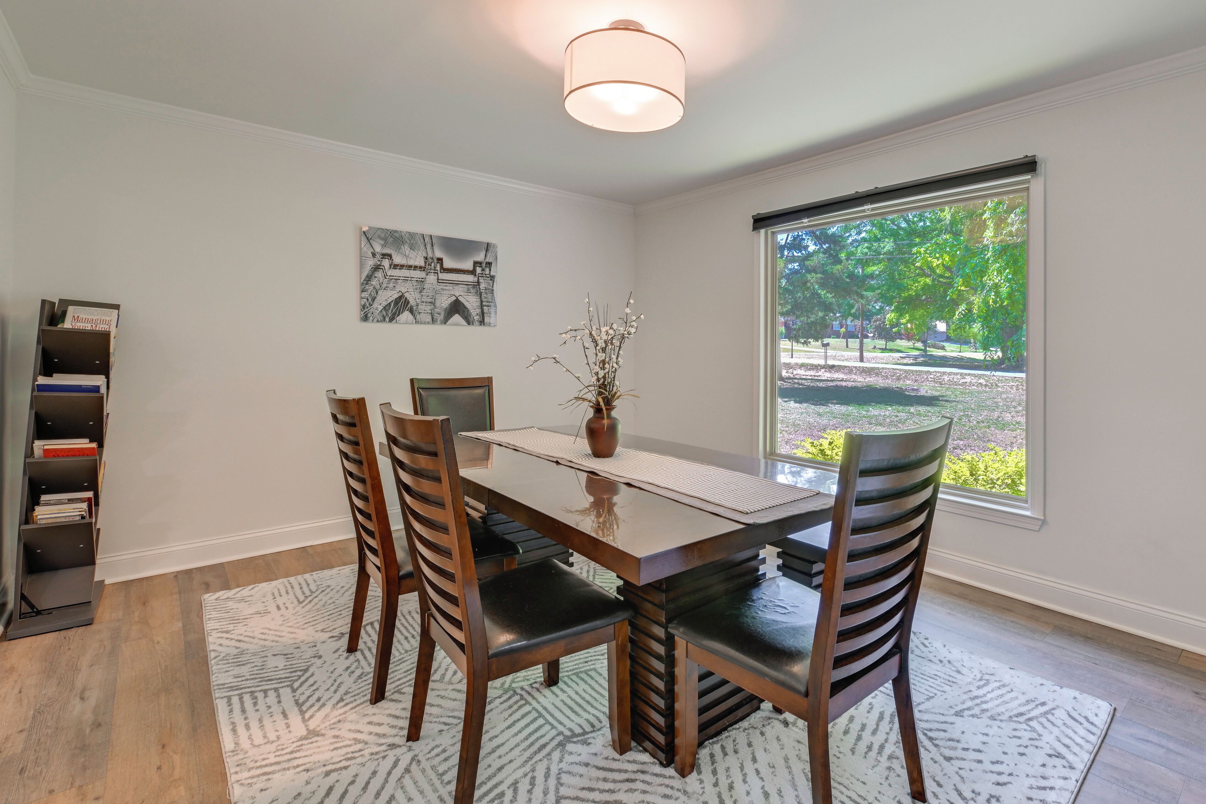 A dining area with a wooden table and chairs, a bookshelf, a framed picture, and a window with a view of trees.