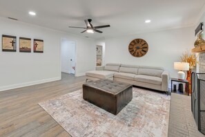 A living room with a beige sectional sofa, a dark brown ottoman, a large wooden clock, and a ceiling fan. - 4 Mi to Charlotte Speedway! Extended Wooded Escape (Harrisburg)