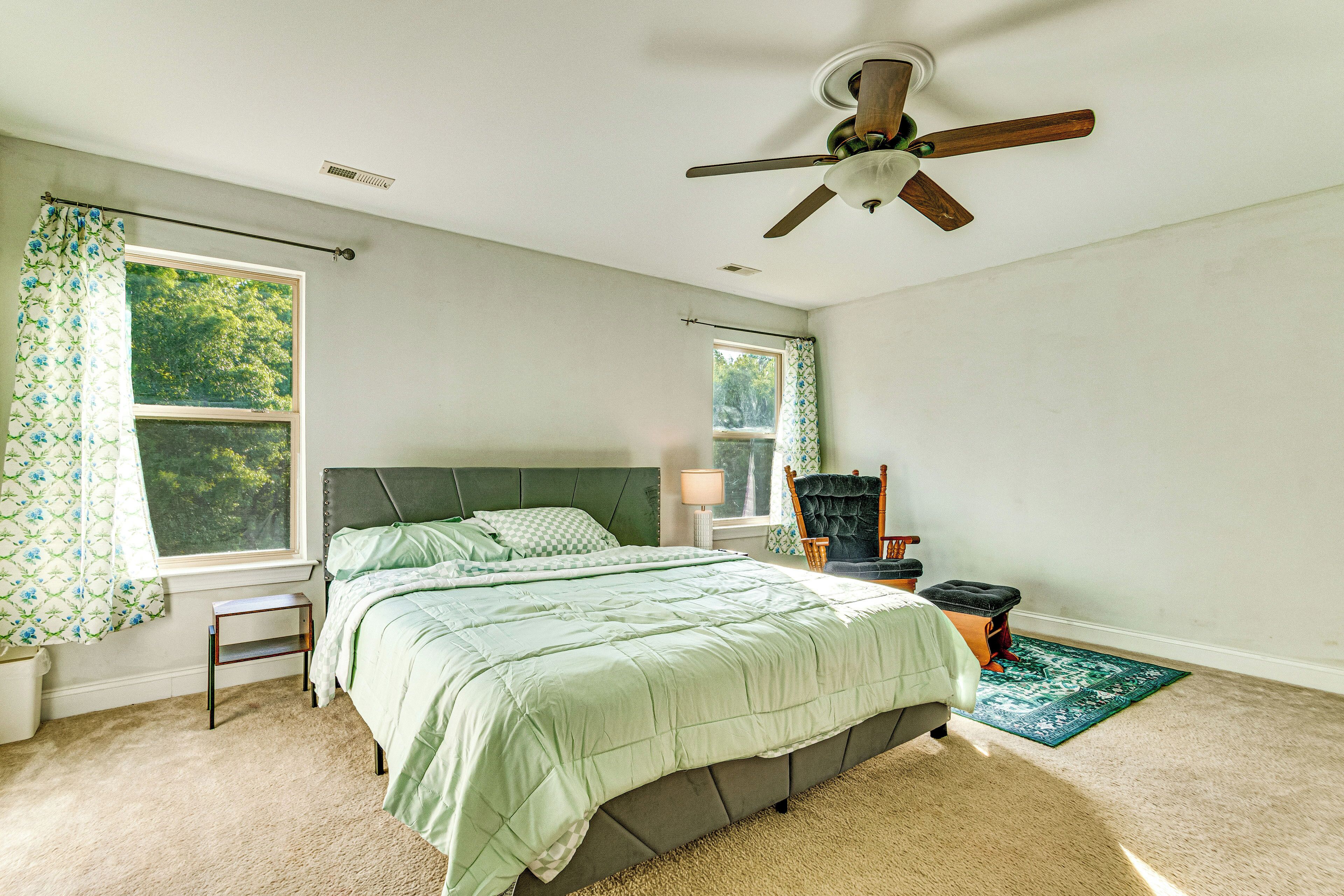 A bedroom with a bed, a ceiling fan, a window with curtains, a nightstand, a chair, and a small table.