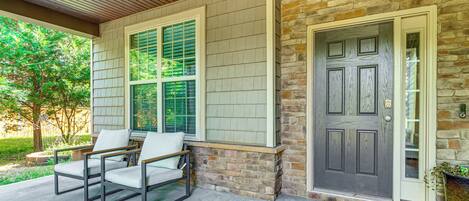 A house exterior with a covered porch, two chairs, a door, and a window with shutters.