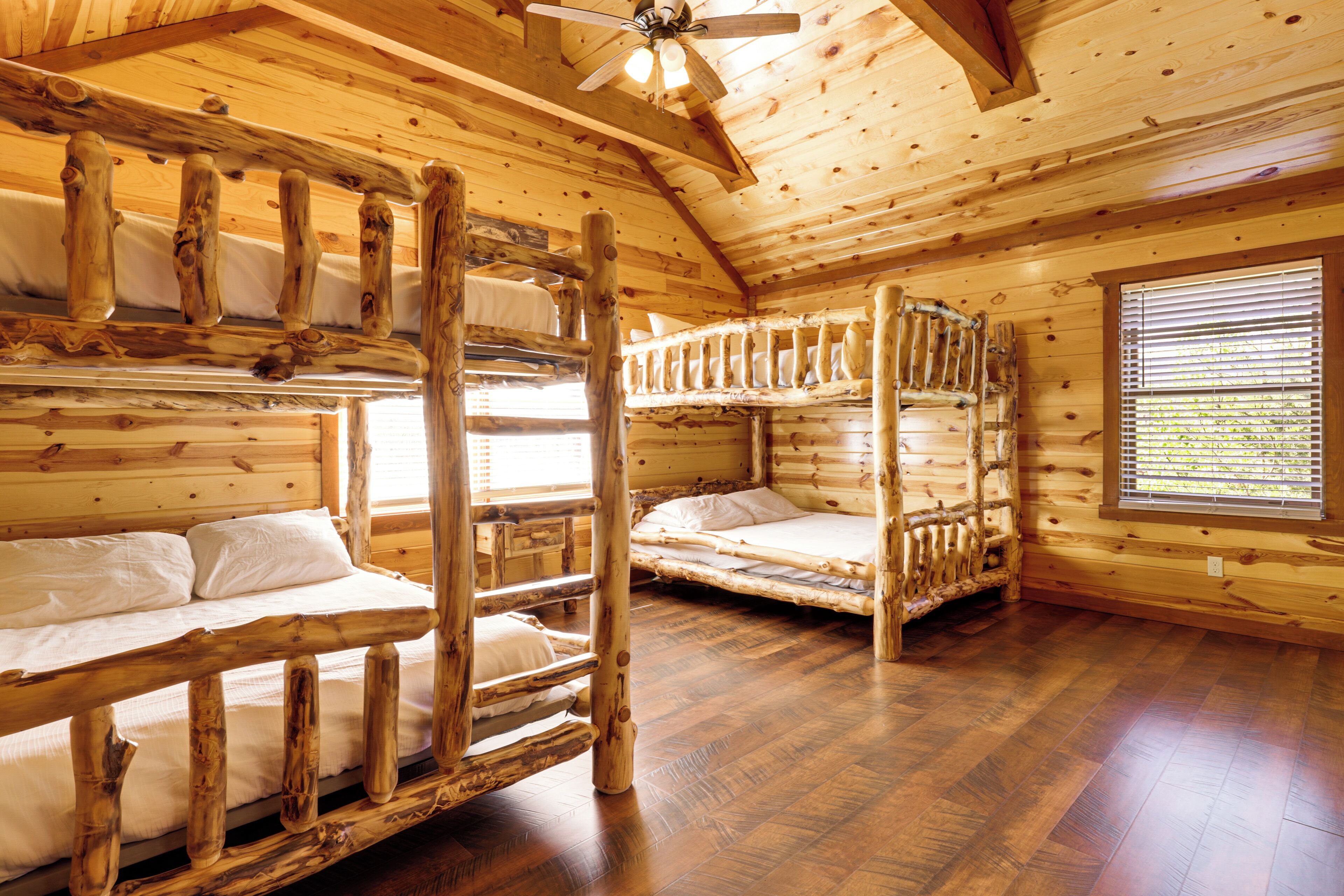 A cabin with wooden walls and floors, bunk beds, a ceiling fan, and a window with blinds.