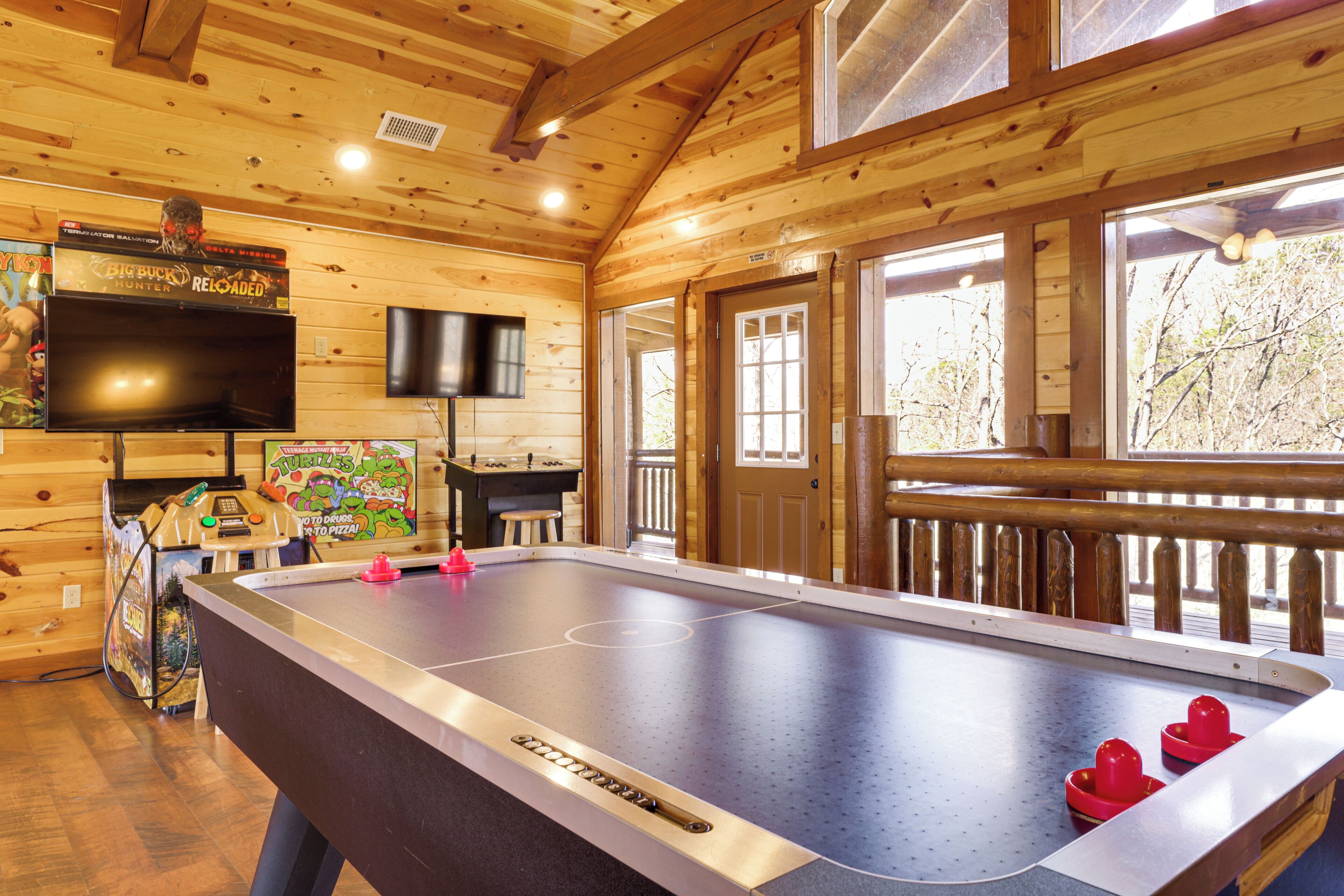 A pool table in a wooden cabin with a television above it and a view of trees outside.