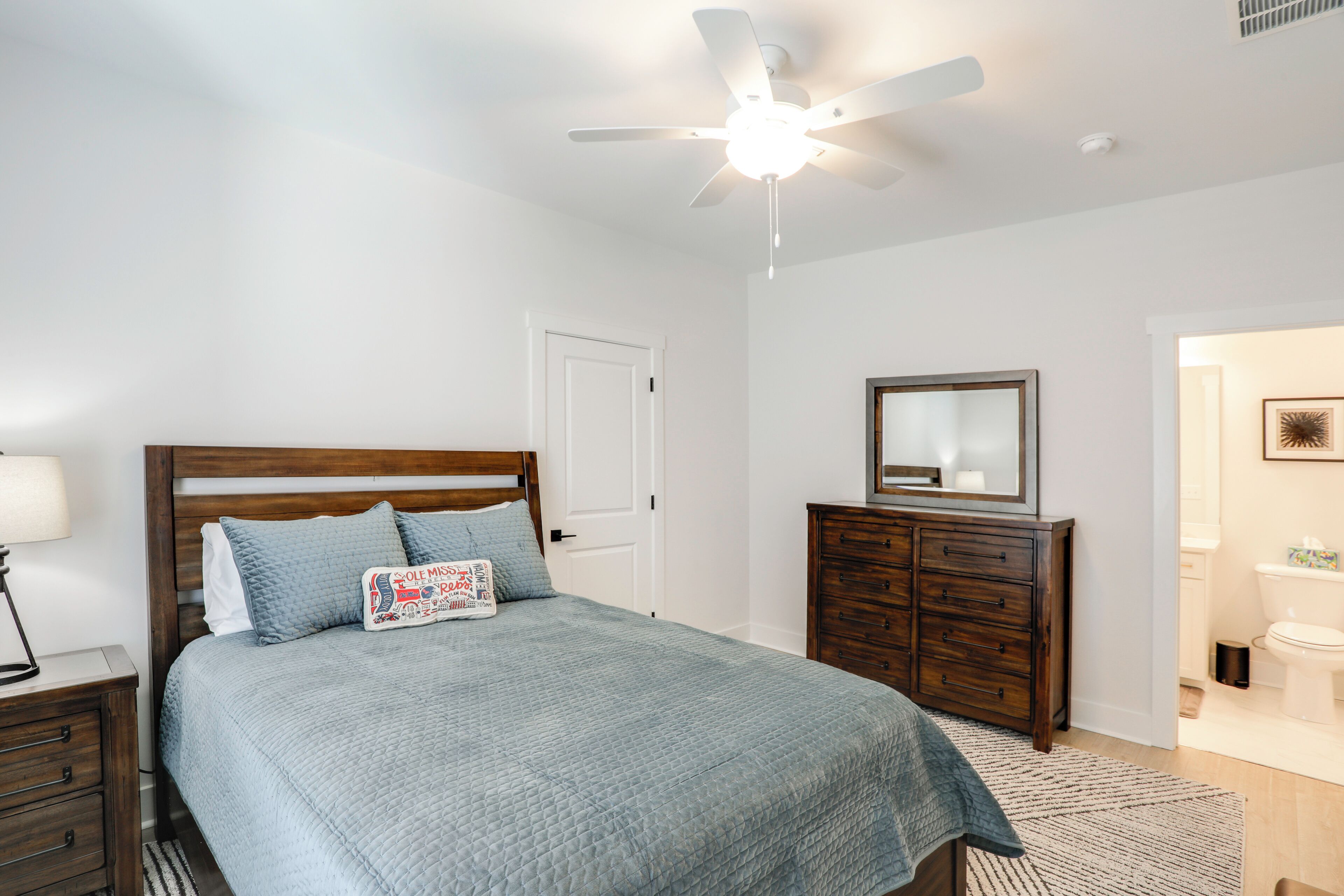 Une chambre à coucher avec un lit, une commode, un ventilateur de plafond et une salle de bain visible à travers une porte ouverte.