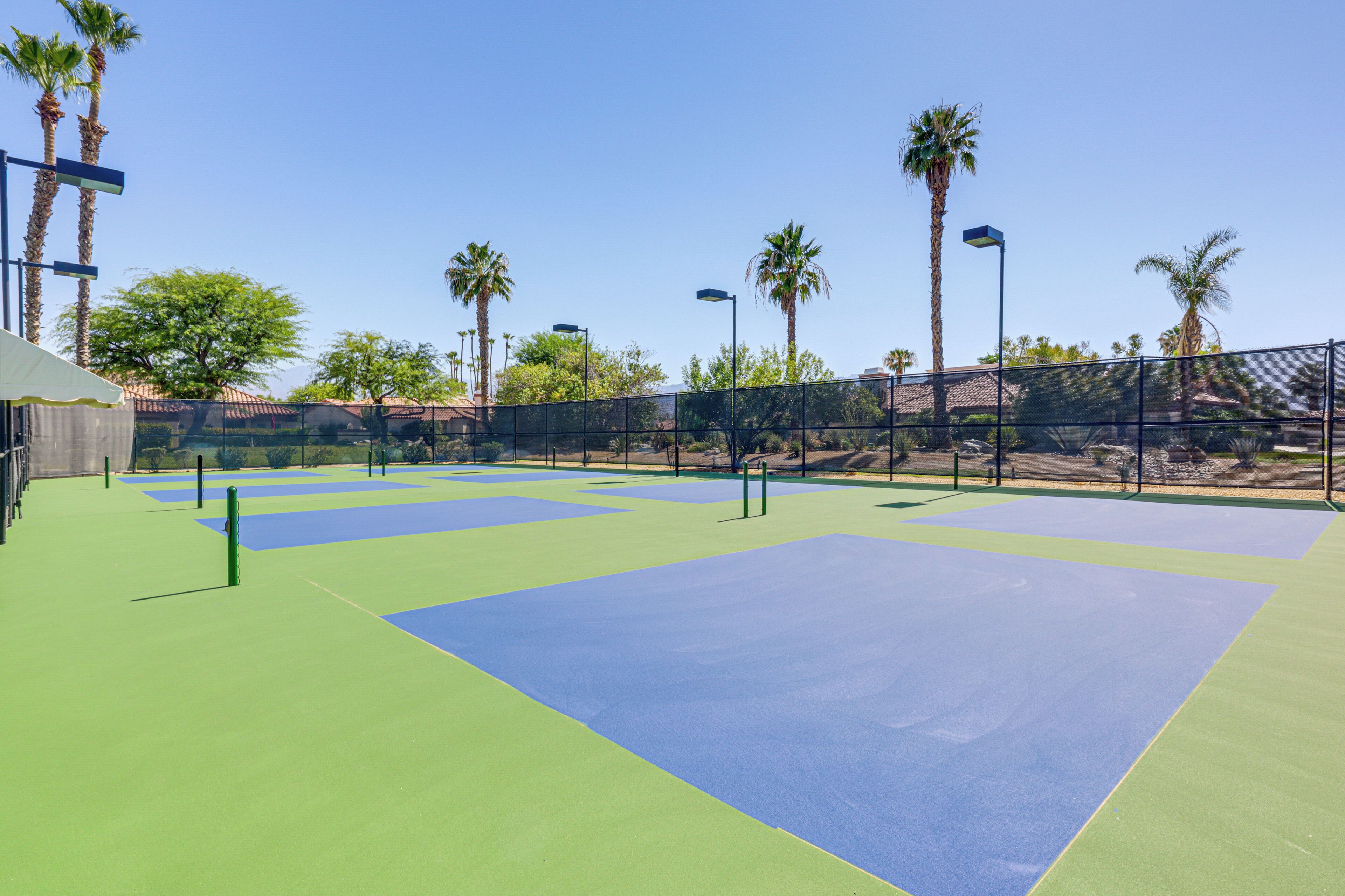 Un court de tennis extérieur aux surfaces vertes et bleues, entouré de palmiers et d’une clôture.