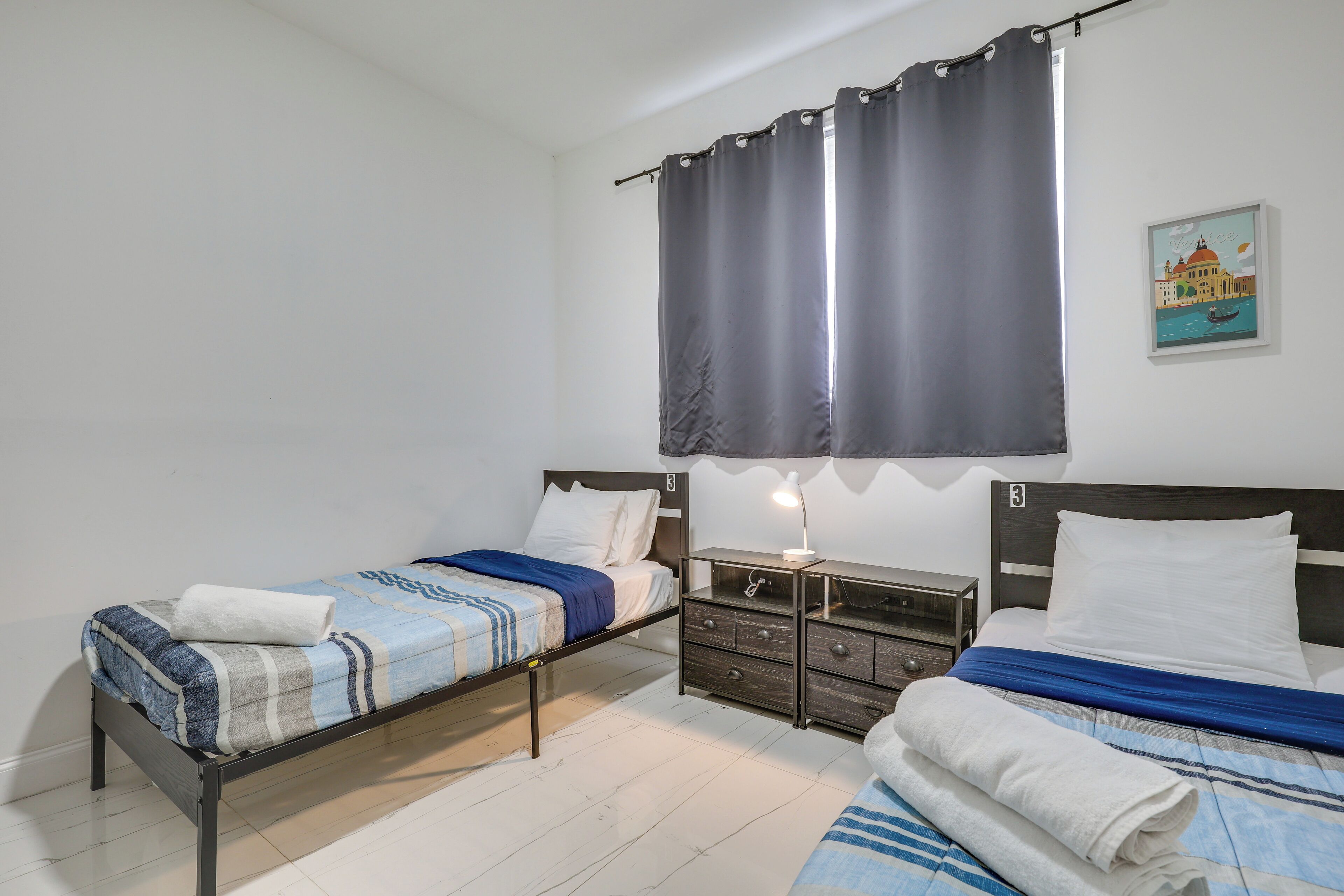 Une chambre avec deux lits simples, une table de chevet, une commode et un rideau.