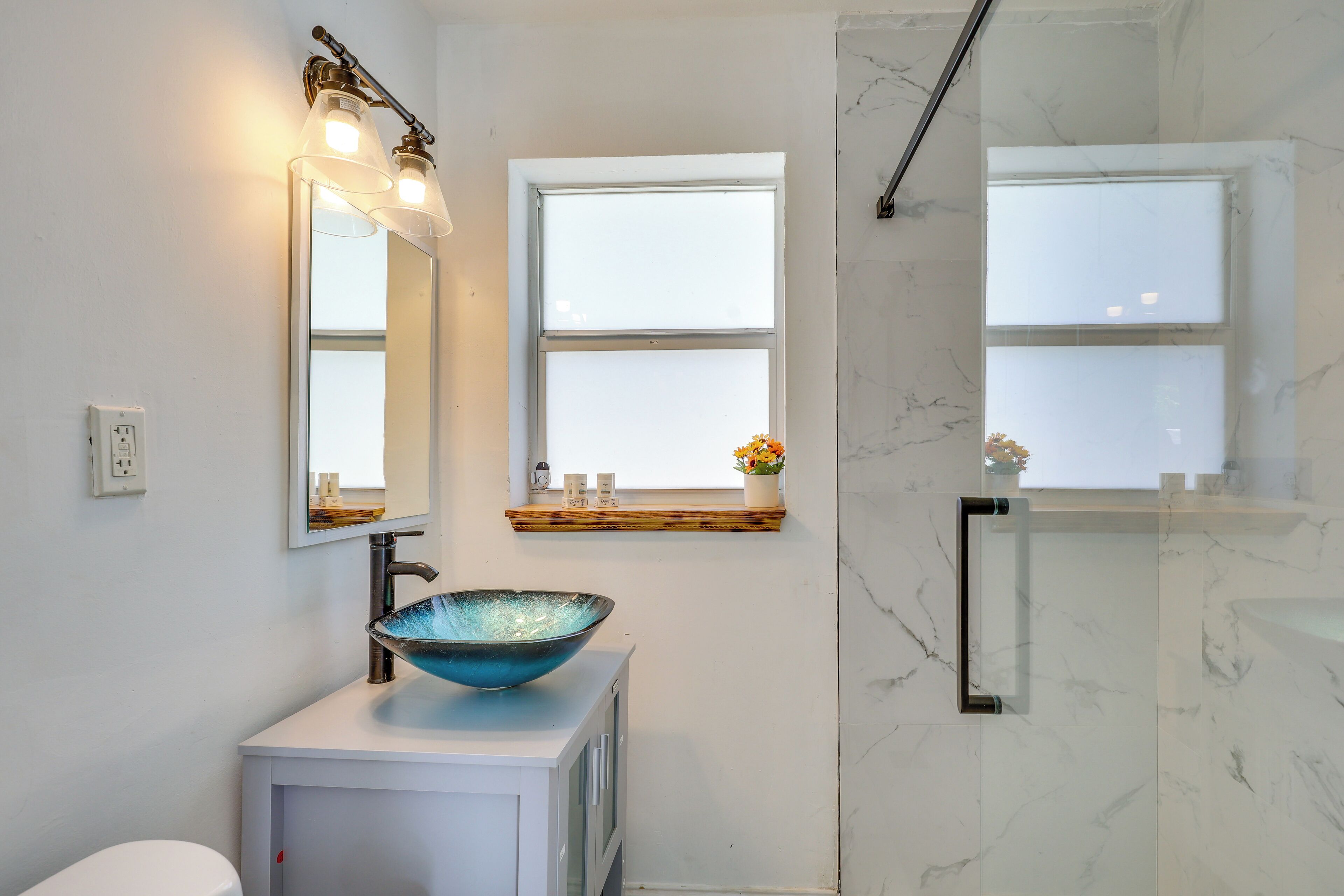 A bathroom with a white sink, a large mirror, and two windows.