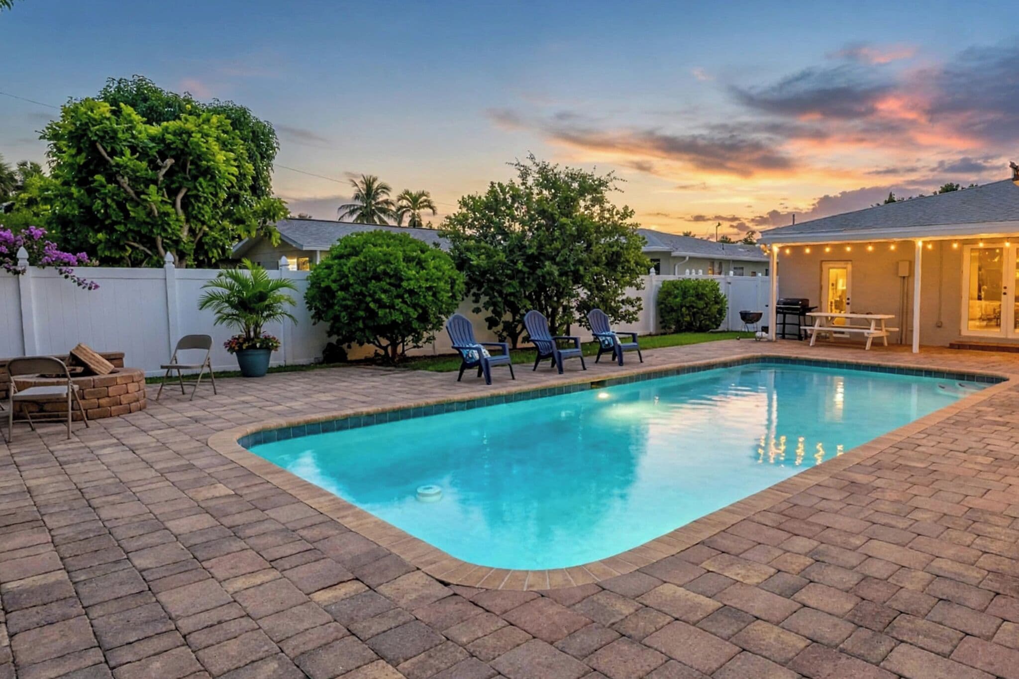 Una piscina rectangular con bordeado de azulejos, rodeada por un patio empedrado. Hay sillas de descanso alineadas a lo largo del borde de la piscina, y se ve una zona cubierta con luces colgantes al fondo.