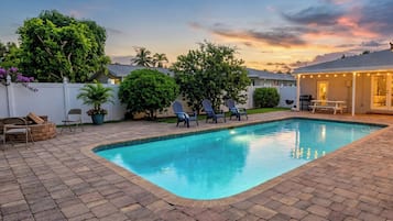 Una piscina rectangular con bordeado de azulejos, rodeada por un patio empedrado. Hay sillas de descanso alineadas a lo largo del borde de la piscina, y se ve una zona cubierta con luces colgantes al fondo.