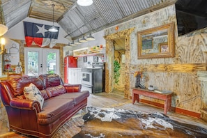 A cozy living room with a leather sofa, a cowhide rug, a Texas flag, and a kitchen area with a refrigerator and stove.
