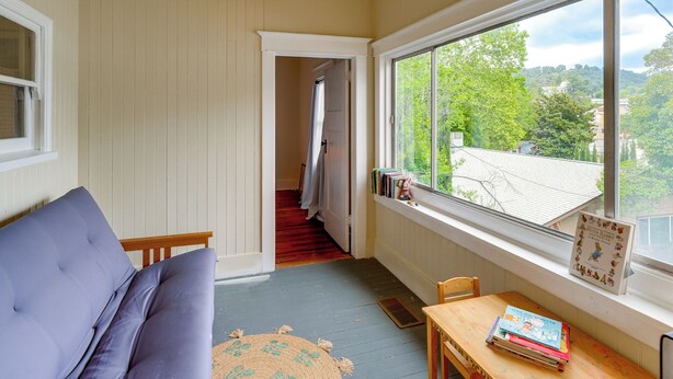 Una habitación con un sofá, una mesa con libros, una ventana con vistas y una puerta que da a otra habitación.
