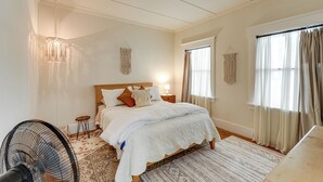 Een slaapkamer met een bed, nachtkastjes, een ventilator en een raam met gordijnen.