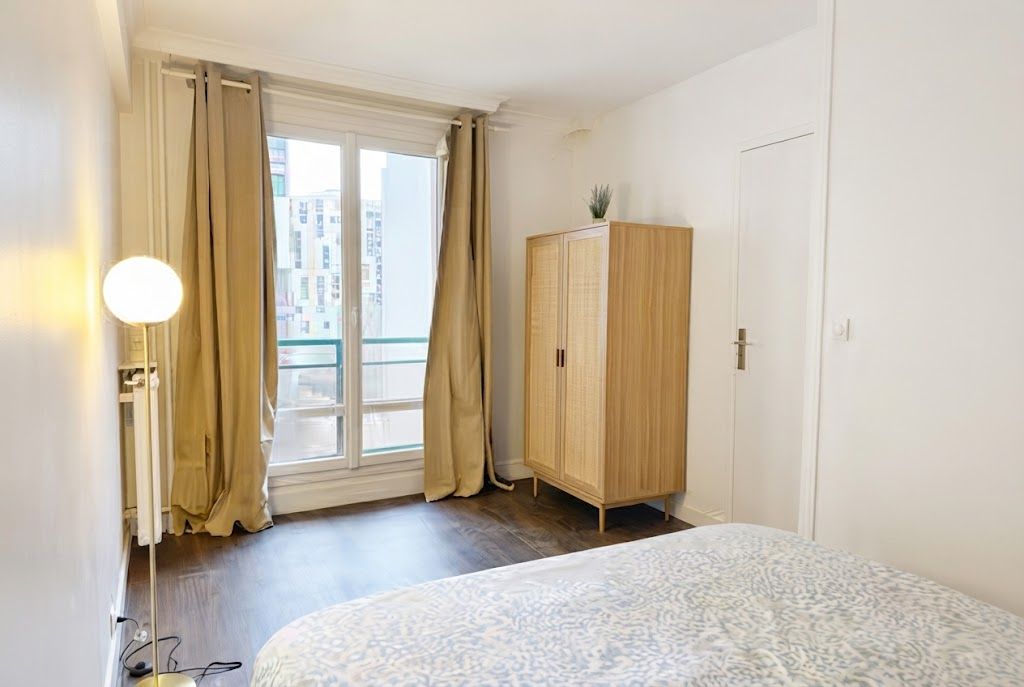 Een slaapkamer met een bed, een kledingkast, een raam met gordijnen en een uitzicht op gebouwen.
