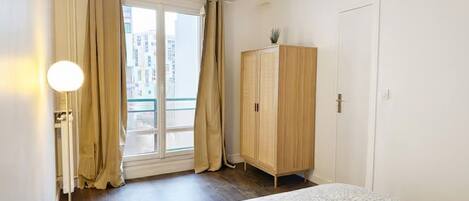 A bedroom with a bed, a wardrobe, a window with curtains, and a view of buildings outside.