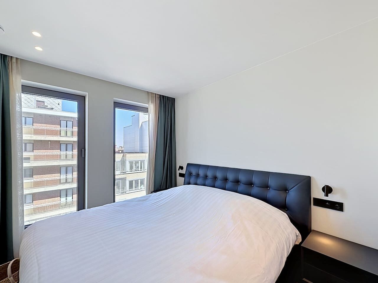 A modern hotel room with a large bed, a nightstand, a wall-mounted control panel, and a view of a building through the window.