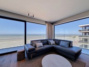 A modern living room with a sectional sofa, a round coffee table, and large windows offering a view of the beach and a high-rise building. - Flat Duin & Dijk - Koksijde (Koksijde)