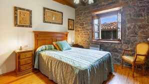 A bedroom with a stone wall, wooden ceiling, a bed with a blue and green bedspread, a wooden nightstand, a chair, and a window with a view of a tiled roof. - Country House 'Casa de Aldea La Quintana' with Mountain Views (Las Viñas)
