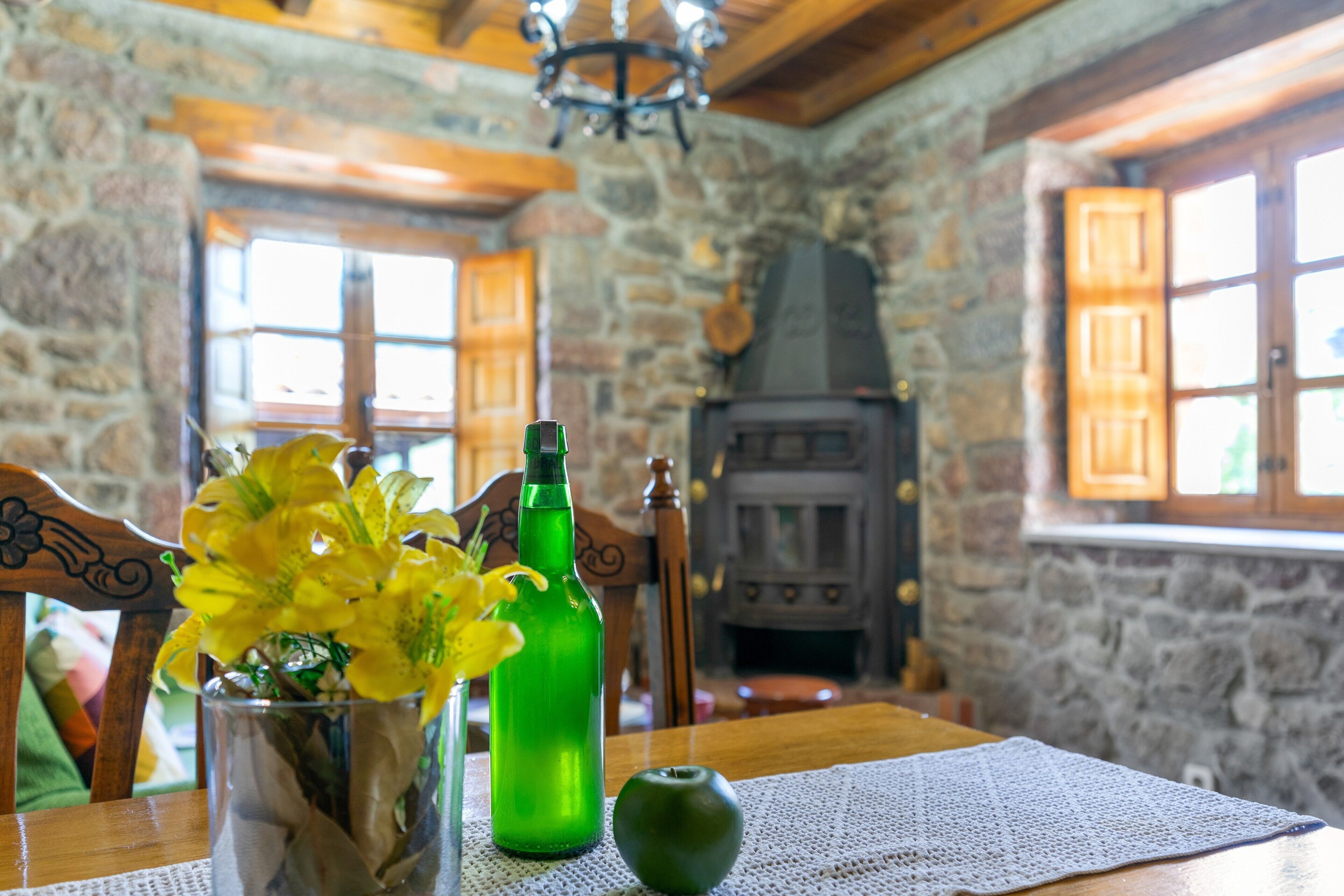 Un interior rústico con pared de piedra, muebles de madera, chimenea y una mesa con un jarrón de flores amarillas.
