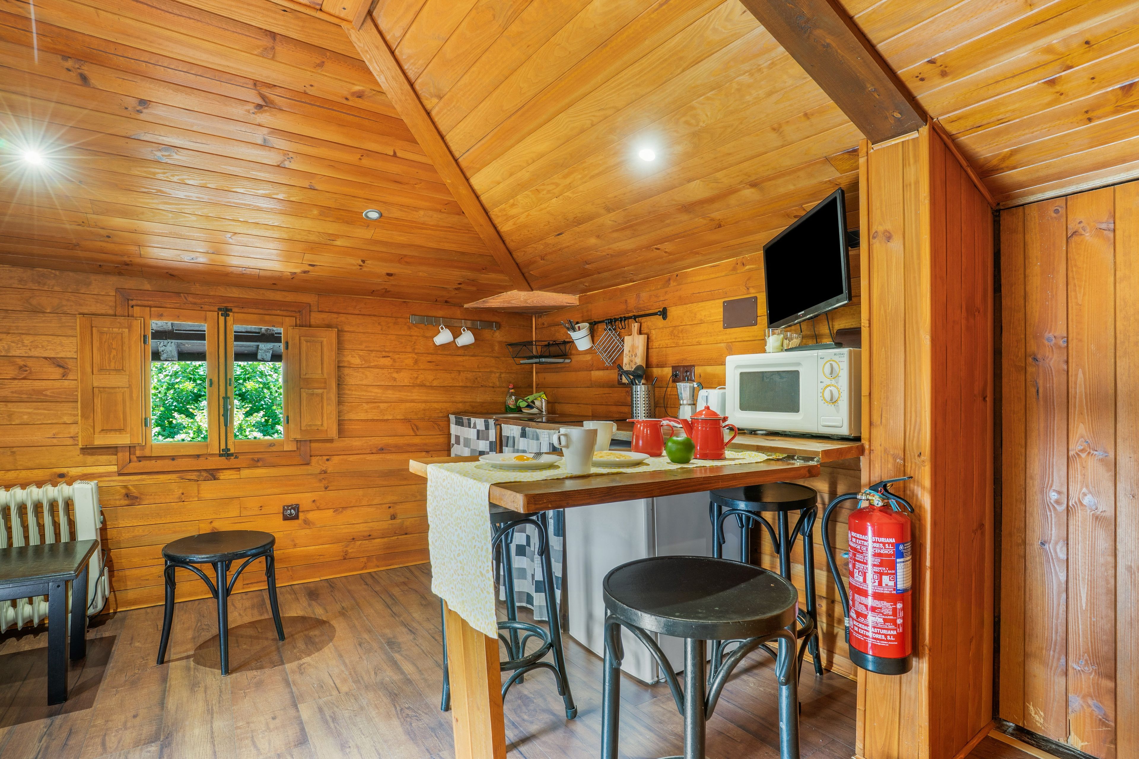 L’intérieur d’une cabane en bois comprend un coin cuisine, deux tabourets, une table, une fenêtre et un extincteur.