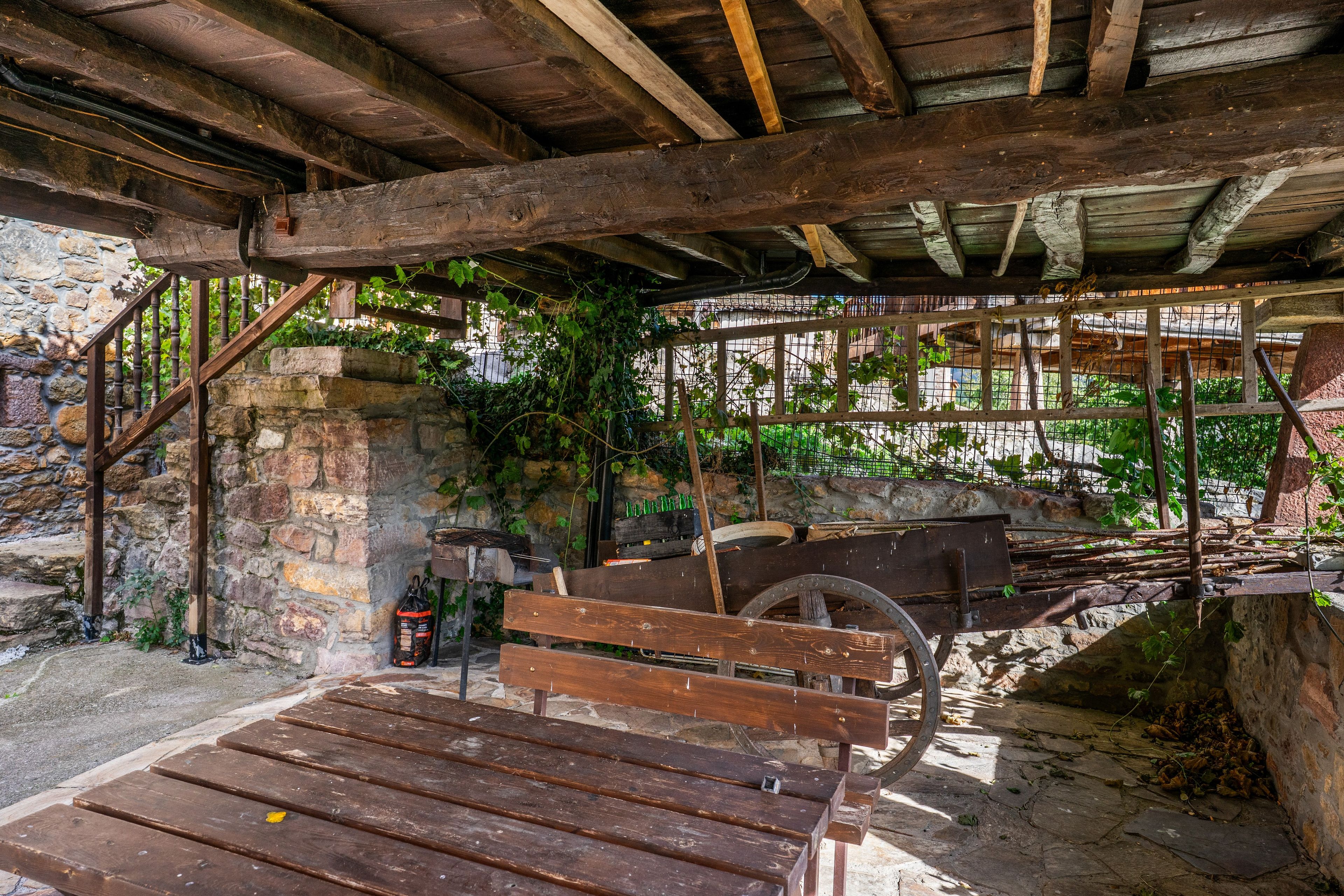 Un área de estar al aire libre rústica con un banco de madera y una estructura circular grande, rodeada de muros de piedra y follaje.