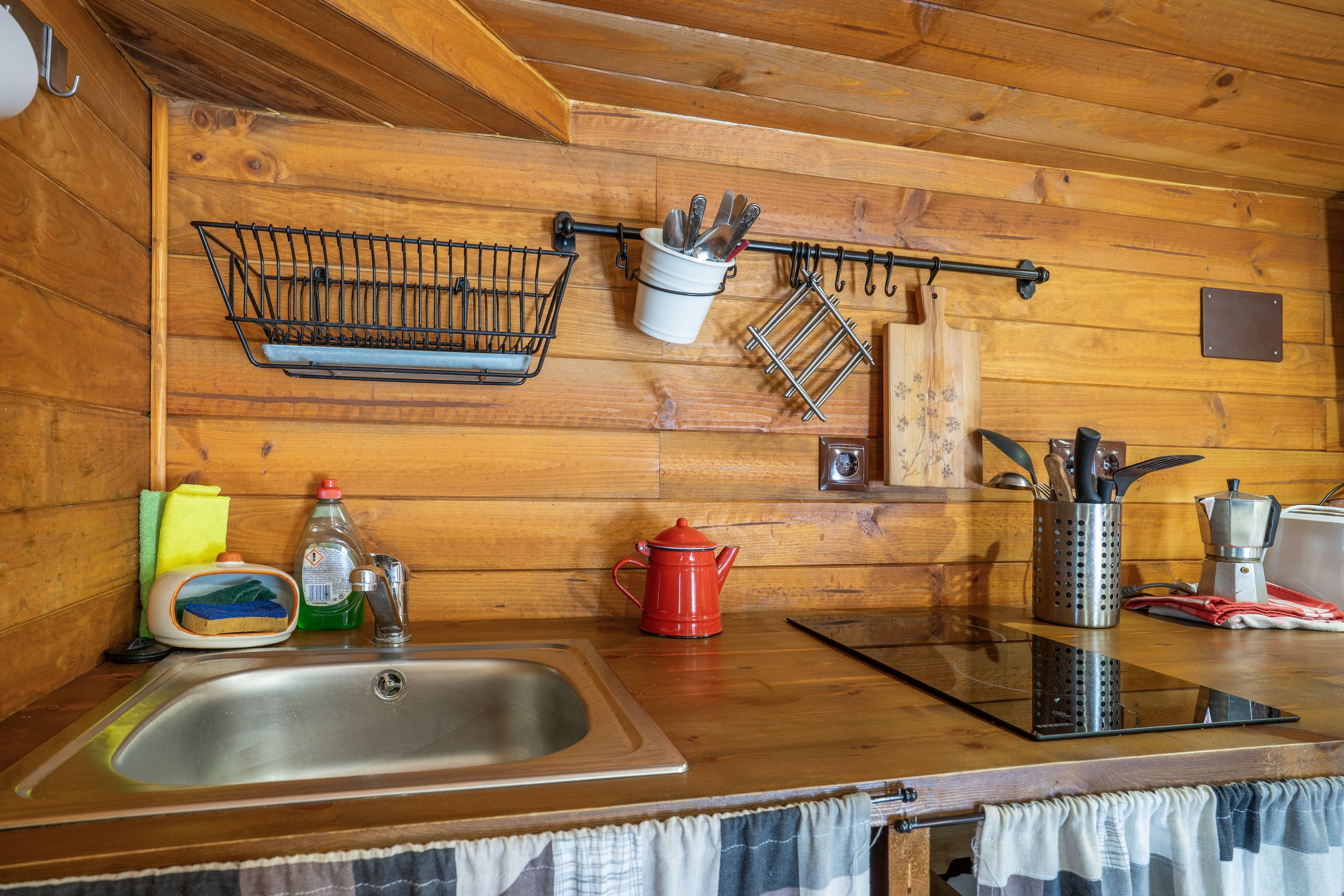 A wooden cabin kitchen with a sink, a red kettle, a coffee pot, and various utensils hanging on the wall.