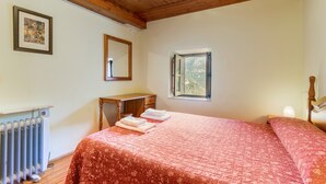 A bedroom with a wooden ceiling, a bed with a floral bedspread, a bedside table, a window with a view of greenery, and a radiator.