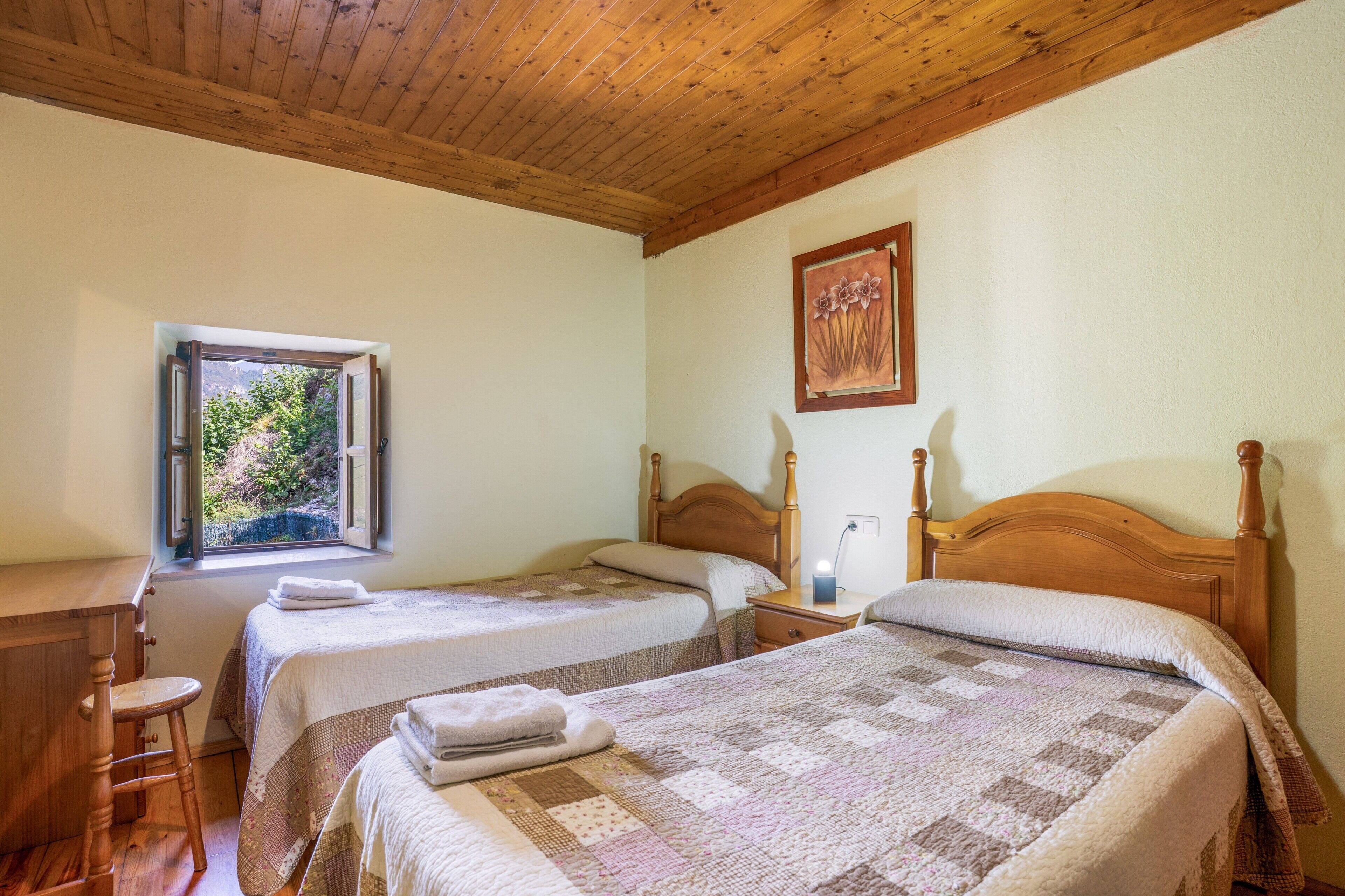Ein Zimmer mit zwei Betten, einem Fenster mit Blick auf Grünflächen, einer Holzbalkendecke und einem Gemälde an der Wand.